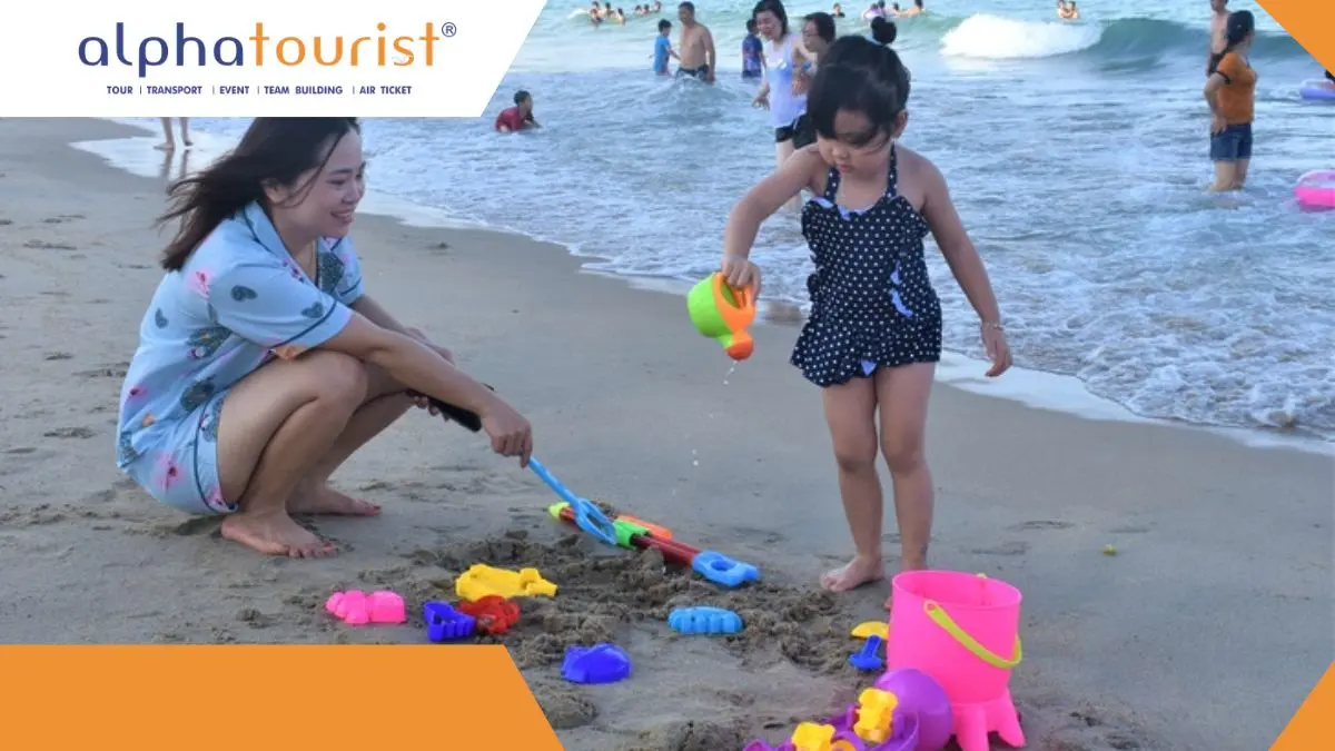 Một em bé Việt Nam khoảng 3 tuổi đang ngồi chơi đồ chơi xúc cát trên bãi biển Bãi Tranh, Nha Trang với làn nước trong xanh và yên tĩnh