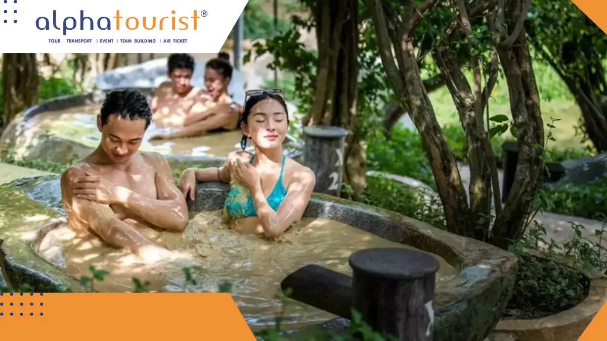 Một cặp đôi đang vui vẻ ngâm mình trong bồn tắm bùn khoáng bằng gỗ, xung quanh là cây cối xanh tươi tại một khu du lịch ở Nha Trang